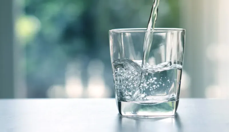 Un jet d'eau fraîche remplit un verre transparent, créant des bulles, avec un fond flou verdoyant.