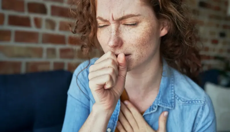 Une femme rousse aux yeux fermés tousse, tenant sa main près de sa bouche et sa poitrine.