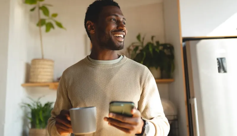 Un homme souriant tient une tasse et un téléphone, regardant au loin dans une cuisine lumineuse.