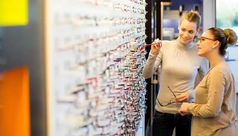 Deux femmes choisissent des lunettes sur un présentoir mural dans un magasin d'optique.
