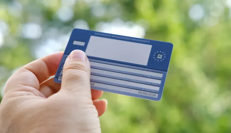 Une main tient une carte européenne d'assurance maladie, sur fond de feuillage vert flou.