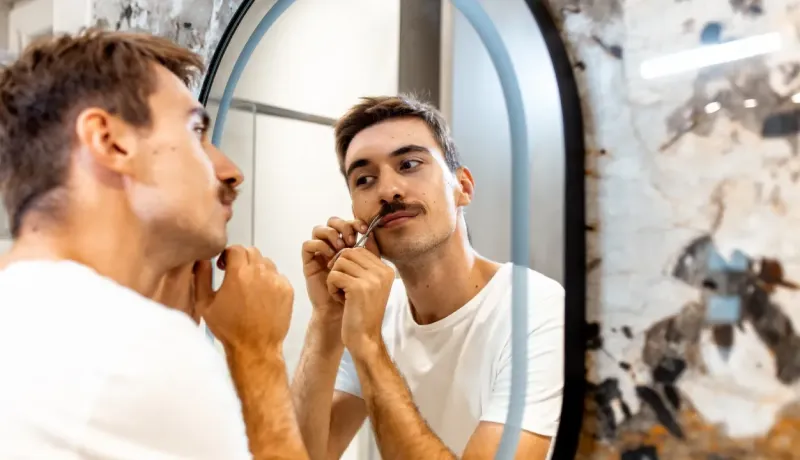 Un jeune homme taille sa moustache devant un miroir rond, avec des ciseaux.