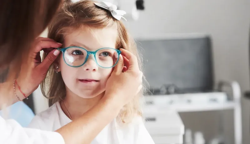 Une adulte ajuste une paire de lunettes bleues sur le visage souriant d'une jeune fille.