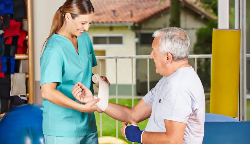 Une physiothérapeute souriante bande le bras d'un homme âgé assis, lui offrant un soutien.