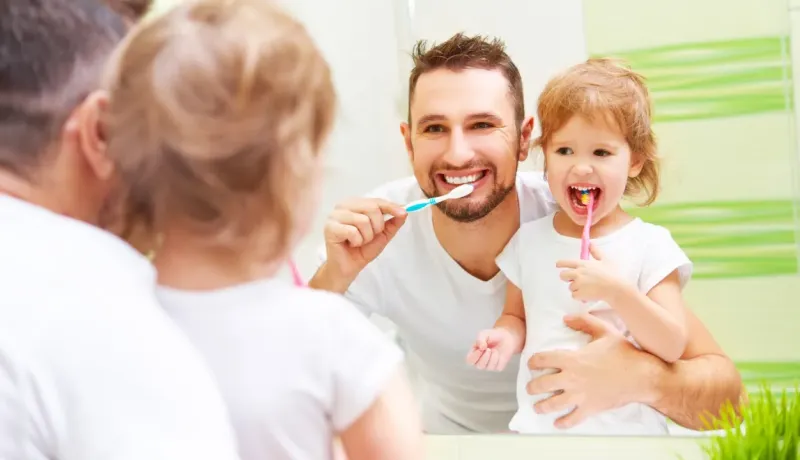 Un père et sa petite fille souriants se brossent les dents ensemble devant un miroir.