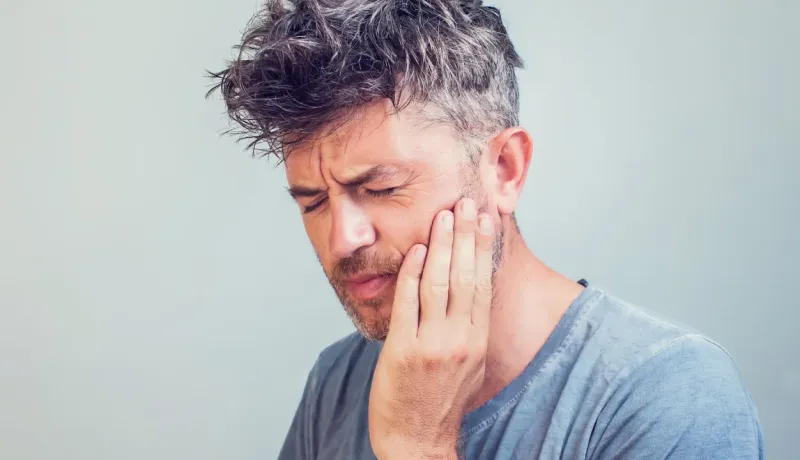 Un homme aux cheveux ébouriffés tenant sa joue, semblant souffrir de douleur dentaire.