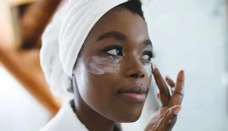 Une femme se met de la crème hydratante sous les yeux, portant une serviette sur la tête.