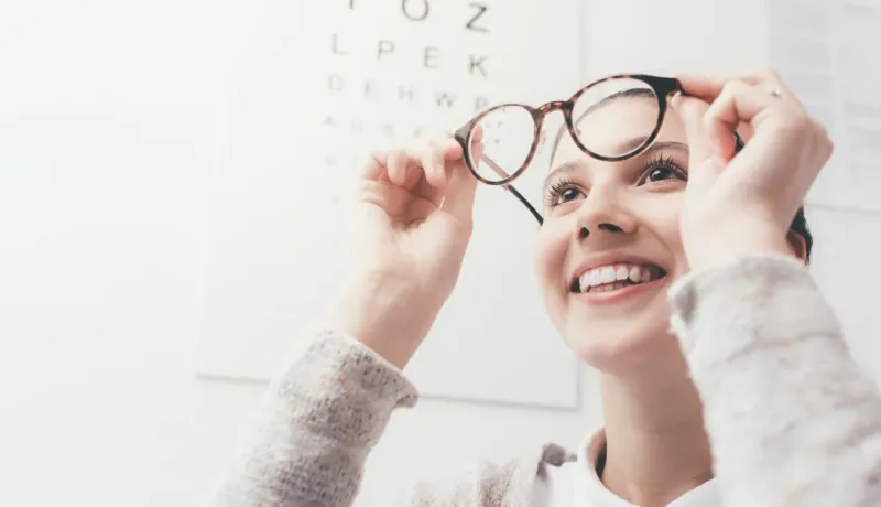 Une jeune femme souriante tient des lunettes devant un tableau optométrique.