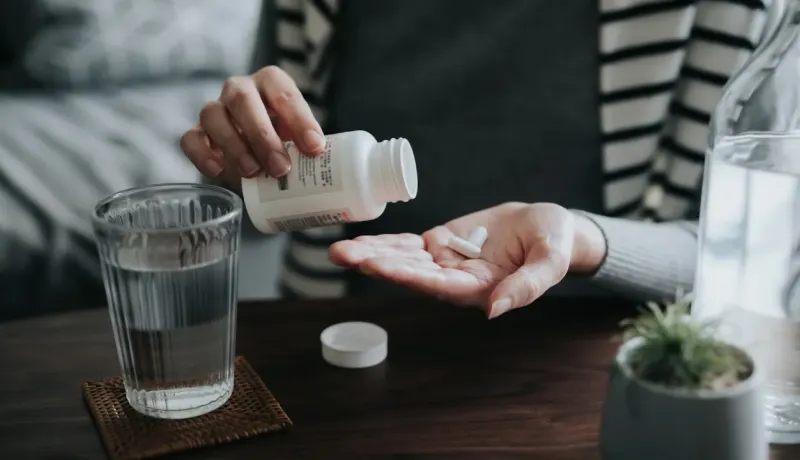 Gros plan sur une personne versant des pilules d'un flacon dans sa main, avec un verre d'eau.
