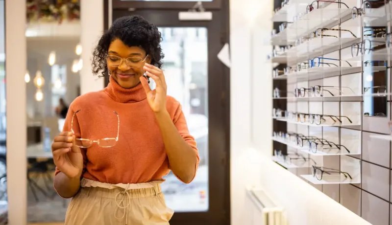 Une femme essaie des lunettes dans un magasin d'optique, souriant à une paire qu'elle tient.