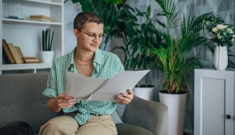 Une jeune femme aux cheveux courts et lunettes lit des documents sur un canapé.