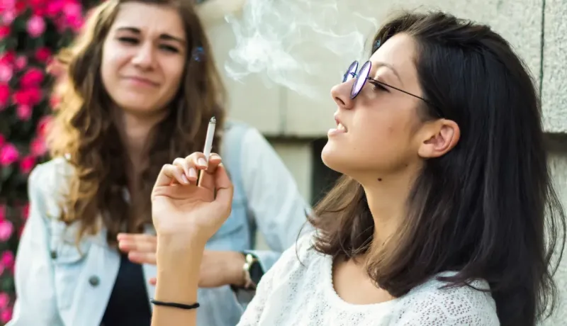 Deux jeunes femmes, l'une fumant et exhalant de la fumée, l'autre en arrière-plan exprimant son dégoût.