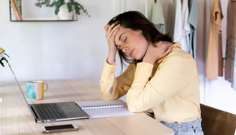 Femme à son bureau, les yeux fermés, se massant le front et le cou, souffrant visiblement d'un mal-être.