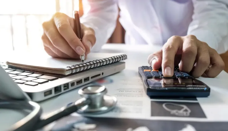 Gros plan sur une personne calculant des chiffres sur une calculatrice, avec un ordinateur portable et un stéthoscope.