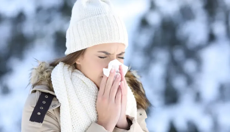 Une femme en tenue d'hiver se mouche dans un mouchoir, l'air souffrant, dans un paysage enneigé.