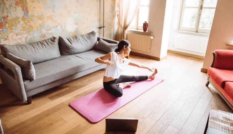 Une femme fait des étirements de yoga sur un tapis rose dans son salon avec son ordinateur portable.