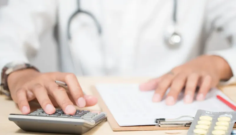 Un médecin calcule sur une calculatrice à son bureau, près de différents documents.