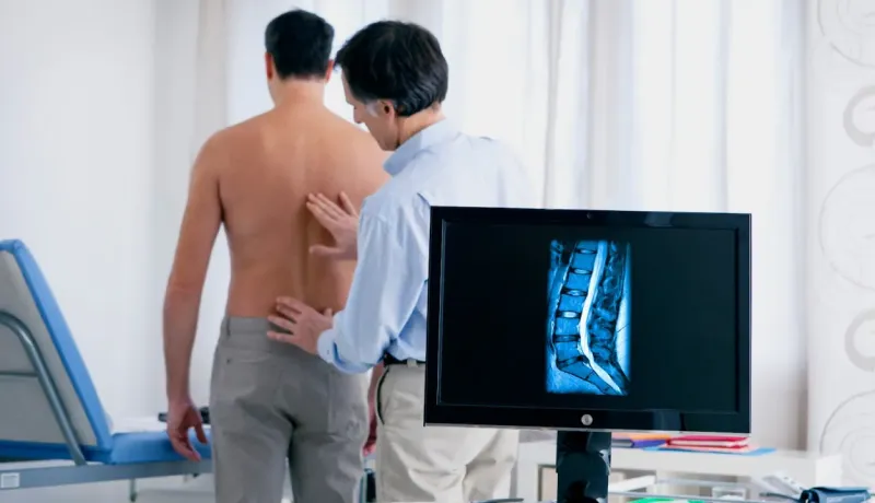 Un médecin examine le dos d'un patient tandis qu'une IRM de colonne vertébrale s'affiche.