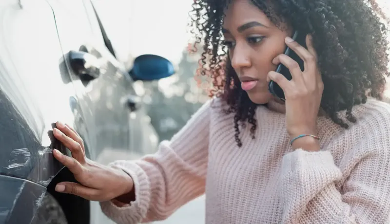 Une femme avec des cheveux bouclés, au téléphone, touche une éraflure sur le côté d'une voiture.