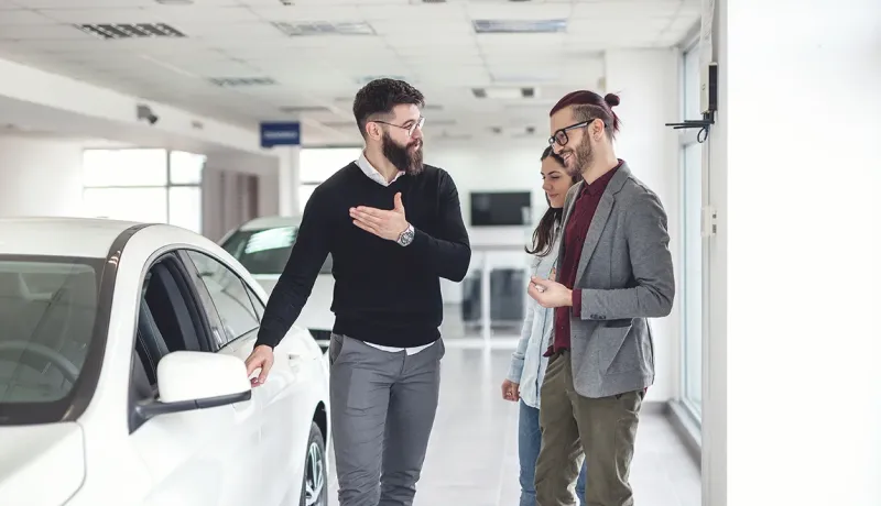 Un vendeur barbu discute avec un couple souriant dans un showroom de voitures, pointant vers une voiture blanche.