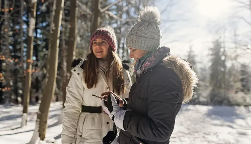 Deux femmes en vêtements d'hiver regardent un téléphone dans une forêt enneigée.