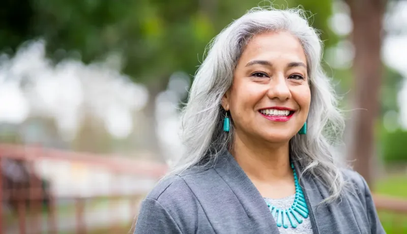 Une femme souriante aux cheveux gris avec des boucles d'oreilles et un collier turquoise, sur fond de verdure.