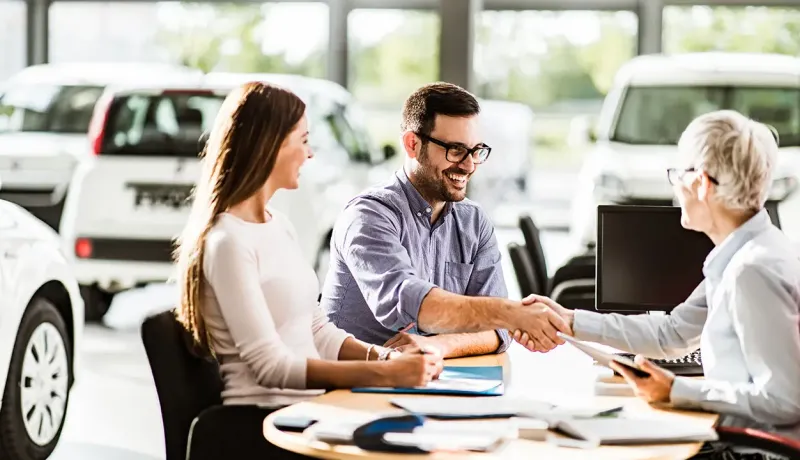 Un couple sourit et un homme serre la main d'une femme à un bureau dans un concessionnaire automobile.