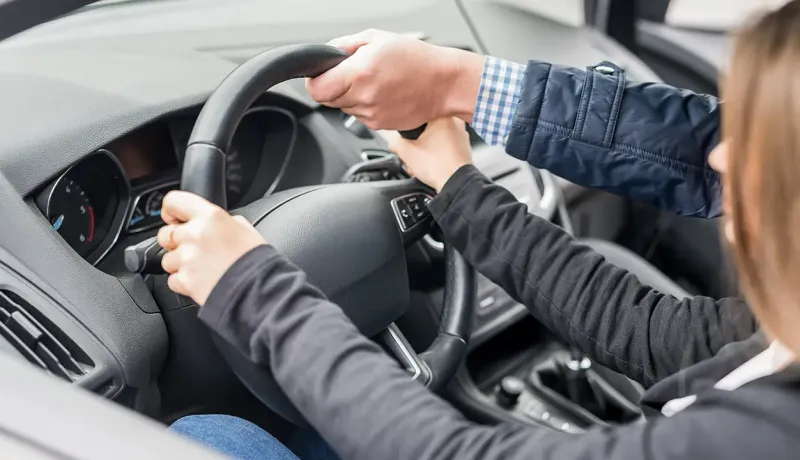 Les mains d'un instructeur guident celles d'un élève sur le volant d'une voiture lors d'une leçon de conduite.