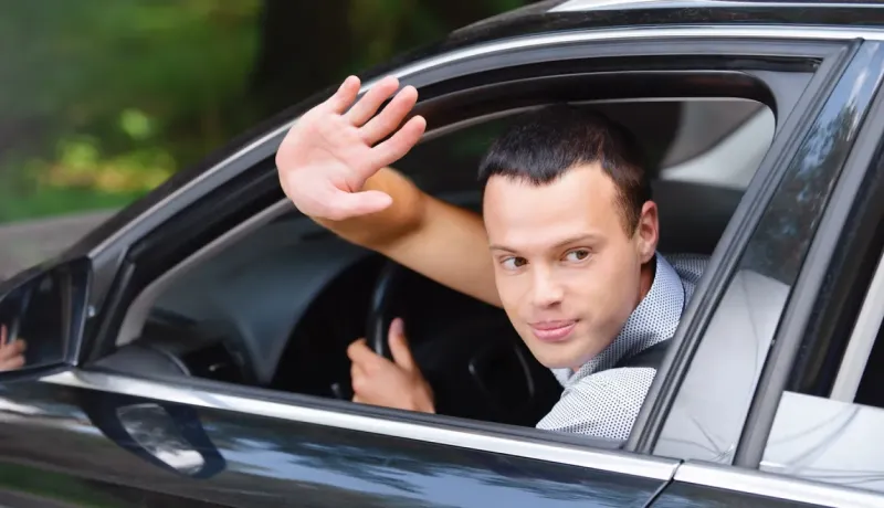 Un homme souriant dans une voiture noire lève la main pour dire au revoir ou saluer, regardant hors de la fenêtre ouverte.