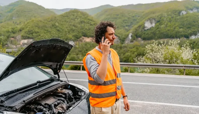 Un homme avec un gilet fluorescent orange est tombé en panne avec son véhicule. Il appelle son assurance pour avoir un dépannage.