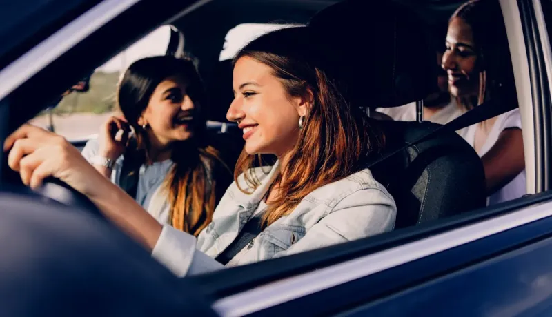Une jeune femme souriante conduit une voiture, avec deux amies assises à l'arrière, profitant du voyage sous le soleil.