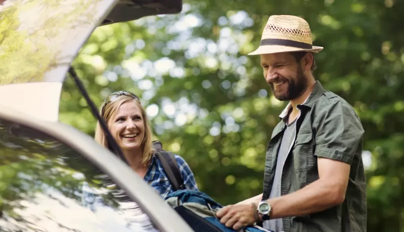 Un couple souriant charge des bagages dans le coffre ouvert d'une voiture.