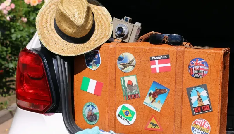 Une valise vintage ornée d'autocollants de voyage est posée à l'arrière d'une voiture, avec un chapeau et une caméra.