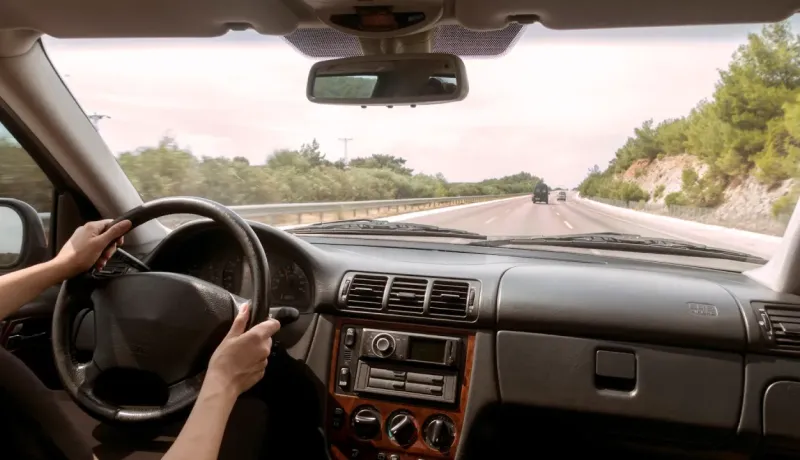 Vue depuis l'intérieur d'une voiture, les mains d'une personne tenant le volant, roulant sur une autoroute bordée d'arbres.