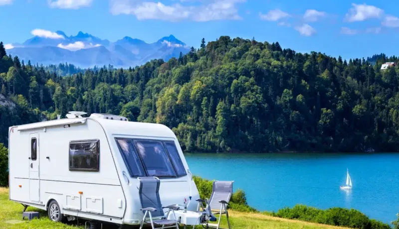 Une caravane blanche est stationnée au bord d'un lac, avec des montagnes et une forêt en arrière-plan sous un ciel bleu.