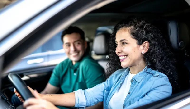 Une jeune femme souriante conduit une voiture, avec un homme souriant sur le siège passager.