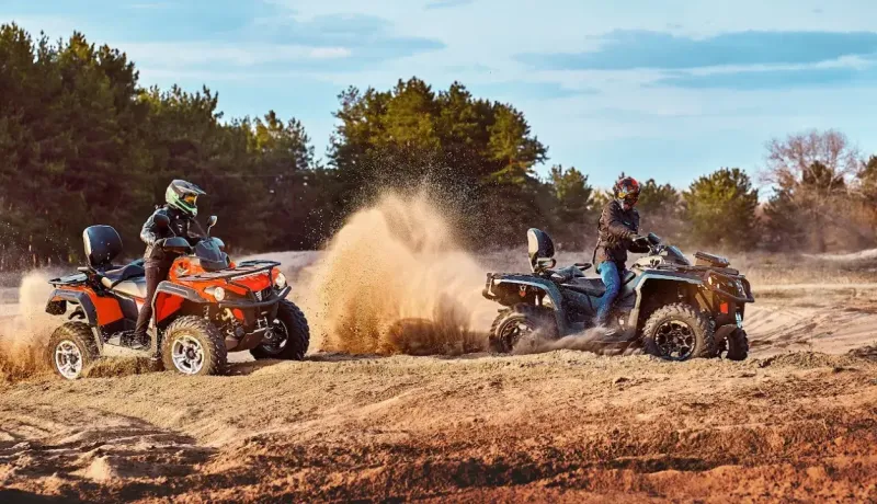 Deux personnes en quads soulèvent de la poussière en roulant sur un terrain sablonneux, avec des arbres en arrière-plan.