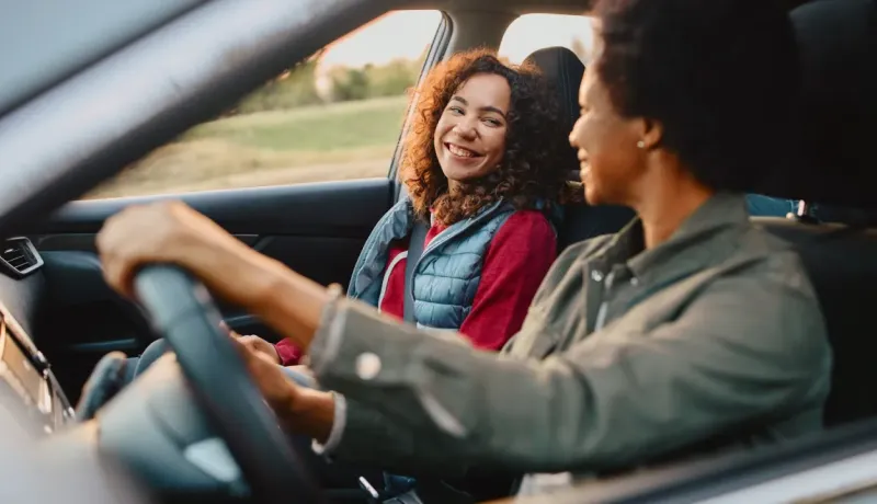 Deux femmes souriantes et heureuses dans une voiture, l'une au volant et l'autre passagère.