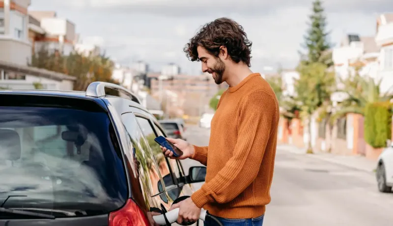 Un homme branche sa voiture électrique chez lui et regarde son téléphone.