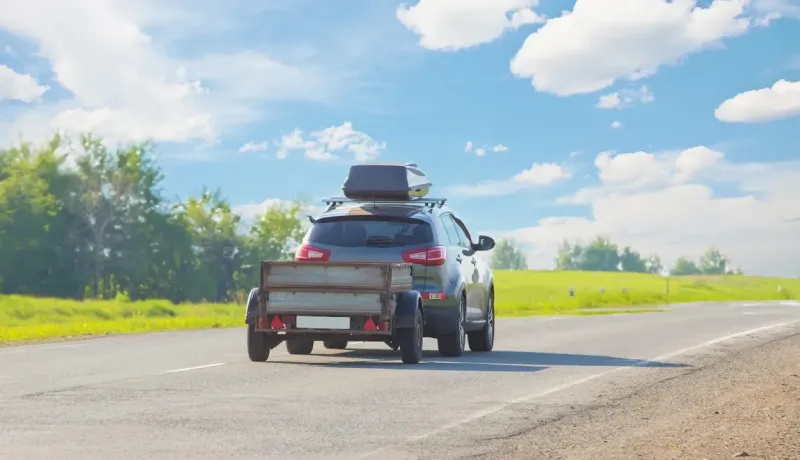 Un SUV gris avec un coffre de toit et une remorque roule sur une route de campagne sous un ciel bleu nuageux.