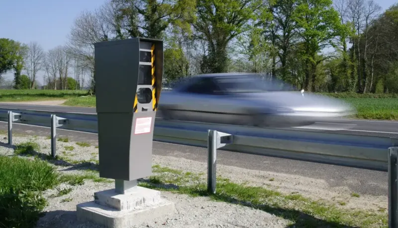 Un radar fixe est installé au bord d'une route, avec une voiture en mouvement rapide et floue en arrière-plan.