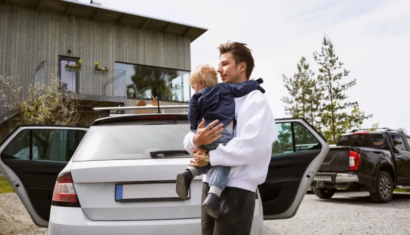 Un homme porte un jeune enfant près d'une voiture familiale blanche avec les portes ouvertes.