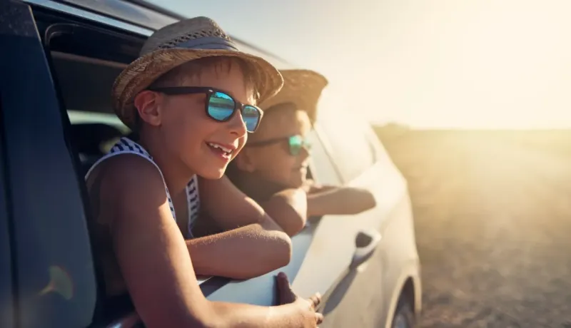 Deux enfants souriants portent des chapeaux de paille et des lunettes de soleil, regardant par la fenêtre d'une voiture.
