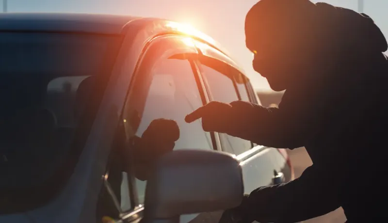 Silhouette d'une personne mal intentionné s'approchant d'une voiture, le soleil couchant créant un fort contre-jour.