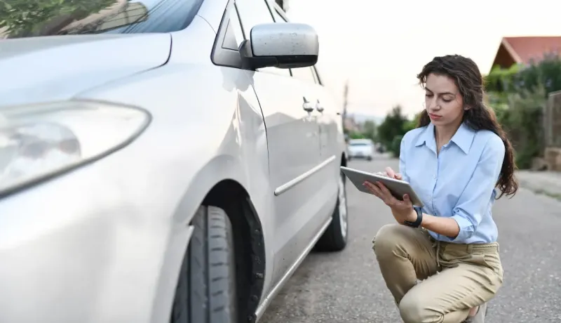 Une femme à genoux à côté d'une voiture, tenant une tablette dans ses mains.