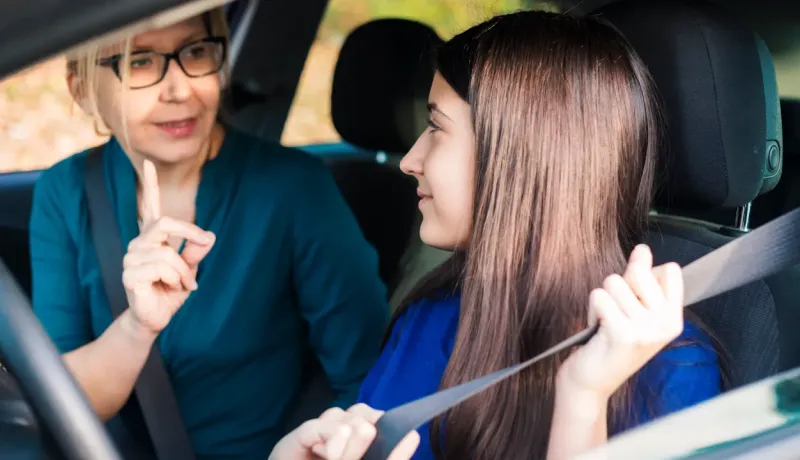 Une mère et sa fille dans une voiture, la mère offrant des conseils à sa fille qui apprend à conduire.