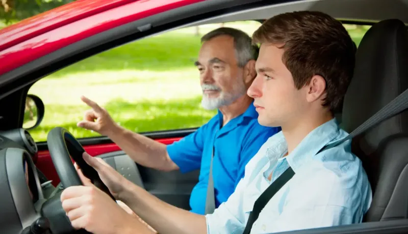Un jeune homme conduit une voiture rouge, un homme plus âgé à ses côtés pointe du doigt, enseignant la conduite.