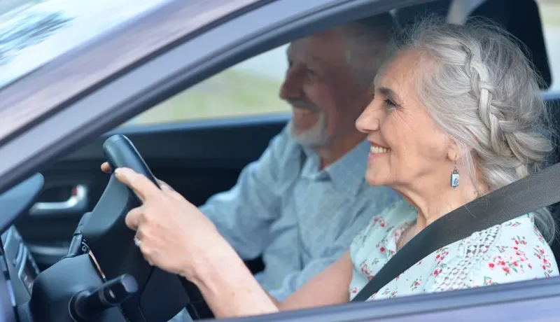 Un couple de personnes âgées souriantes est assis dans une voiture, la femme tient le volant.