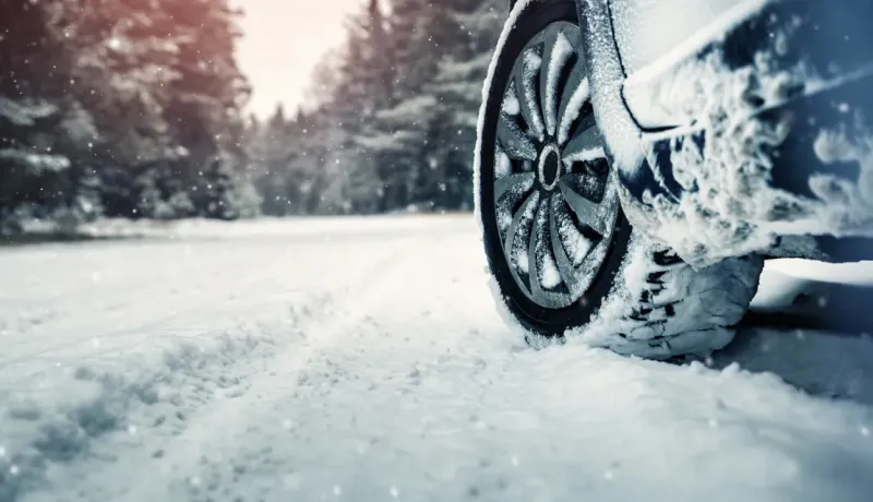 Un pneu de voiture enneigé est visible sur une route couverte de neige, avec des arbres en arrière-plan.