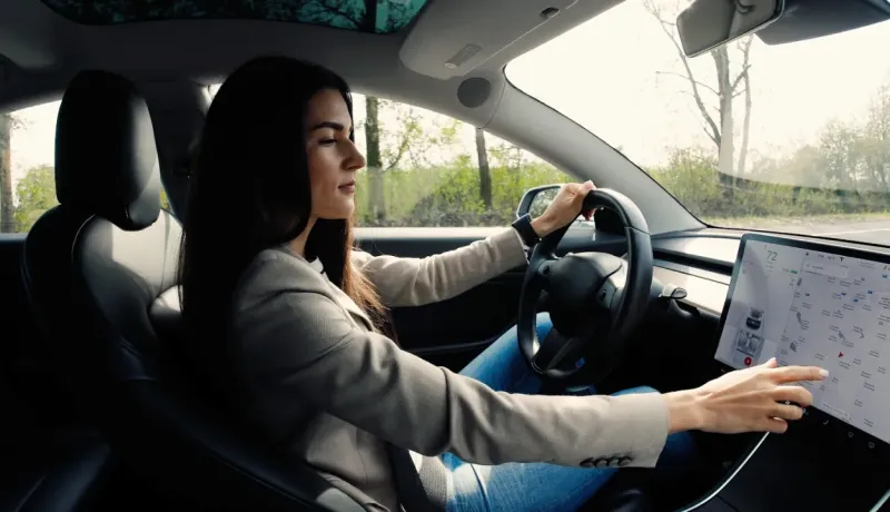 Une jeune femme aux cheveux longs conduit une voiture connectée, son regard tourné vers le tableau de bord numérique.
